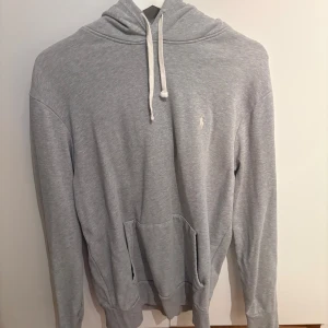 Grå hoodie från Polo Ralph Lauren - Snygg ljusgrå hoodie från Polo Ralph Lauren med klassisk huva och vit snörning. Tröjan har en broderad logga på bröstet, känguruficka och är tillverkad i mjuk bomullsmix. Perfekt för chill dagar eller när du vill ha en clean look.