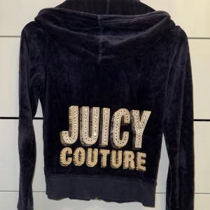 Vintage Juicy Couture Mörk marinblå velour hoodie - Svart velour hoodie från Juicy Couture med glittrande guldfärgade stenar och stor JUICY COUTURE-text på ryggen. Tröjan har huva, dragkedja framtill och fickor. Några få sprickor i tyget men inget som syns på längre håll.