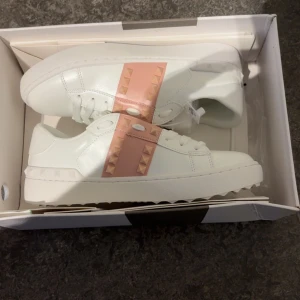 Valentino Rockstud sneakers vit/rosa - Snygga Valentino Garavani Rockstud sneakers i vitt skinn med bred rosa rem över vristen dekorerad med ikoniska nitar. Klassisk rund tå, platt sula och vita snören. Perfekt för dig som vill sticka ut med exklusiva detaljer och modern design.