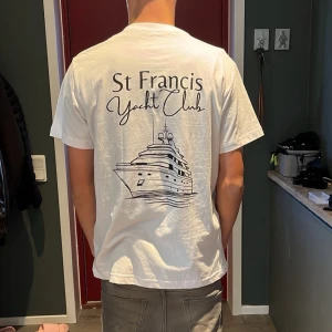 Vit St Francis Yacht Club t-shirt - Vit t-shirt med tryck av St Francis Yacht Club och en stor yacht på ryggen. Klassisk passform med rund hals och korta ärmar. Skön och luftig i bomull, perfekt för chill dagar eller när du vill ha en clean stil.