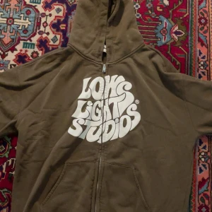 Brun hoodie från Lowlights Studios M - Brun hoodie från Lowlights Studios i storlek medium med stor vit grafisk logga på framsidan. Tröjan har huva, dragkedja och två fickor framtill. Mjuk insida och klassisk streetwear-look, perfekt för chill dagar.