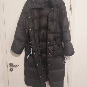 Svart lång pufferjacka med huva - Säljer en svart lång pufferjacka med huva och dragkedja. Jackan har quiltad design, långa ärmar och reflexdetaljer på insidan. Perfekt för kalla dagar och riktigt skön att bära. Stängs med både dragkedja och knappar.Helt ny.Har tagit bort ettiket.