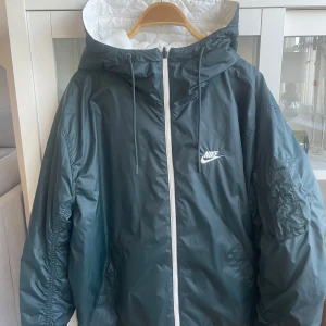 Nike vindjacka med huva och dragkedja - Nike vindjacka med huva, hel dragkedja och snörning. Två sidig jacka.    Jackan är färgblockad i vitt, mörkgrönt och olivgrönt med broderad Nike-logga på bröstet. Lättviktig och loose fit, perfekt för blåsiga dagar. Flera fickor och cool streetstil