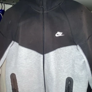 Nike hoodie i svart och grå med luva  - Nike hoodie i tvåfärgad design med svart upptill och ljusgrå nedtill. Tröjan har hel dragkedja, huva och två fickor med dragkedja. Nike-logga i vitt på bröstet. Perfekt för chill eller träning.
