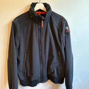 Svart softshelljacka Parajumpers L - Säljer min Parajumpers Soft Shell-jacka i storlek L. Den är i nyskick och har endast använts sparsamt. Jackan har en dold huva som enkelt kan tas fram eller stoppas undan med dragkedjan i nacken. Jag är 184 cm lång och den sitter perfekt på mig. Färgen är mörkblå med snygga orangea detaljer, två framfickor med dragkedja och Parajumpers klassiska logga på ärmen.