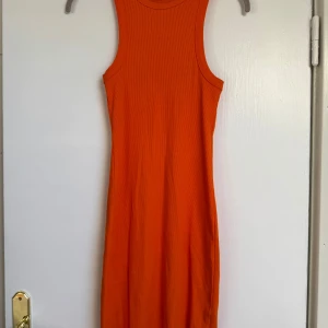 Ribbad orange ärmlös klänning - Säljer en snygg orange ribbad klänning med ärmlös design och rund hals. Klänningen har en kroppsnära passform och är perfekt för dig som gillar färgstarka plagg. Materialet är mjukt och stretchigt, vilket gör den bekväm att bära hela dagen.