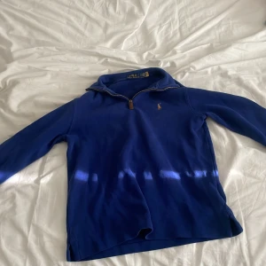 Blå half zip tröja från Polo Ralph Lauren - Säljer en blå långärmad tröja från Polo Ralph Lauren med half zip och klassisk broderad logga på bröstet. Tröjan har hög krage och är i mjukt bomullsmaterial, perfekt för lager-på-lager. Enkel och stilren design som funkar året runt.