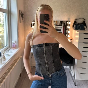 Svart denim korsett-topp från Zara - Säljer en svart, croppad korsett-topp från Zara i denim med markerade sömmar och dragkedja framtill. Toppen är axelbandslös och har smockad rygg för extra komfort. Perfekt att styla med jeans eller kjol för en cool look. Använd endast några fåtal gånger 🖤