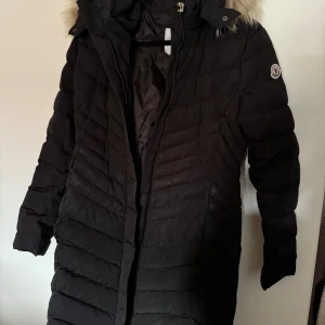 Svart lång dunjacka från Moncler - Säljer en svart lång dunjacka från Moncler med huva och snygg pälsdetalj. Jackan har quiltad design, dragkedja framtill och Moncler-logga på ärmen. Perfekt för kalla dagar och riktigt stilren look. Dragkedjan har hängt sig men går enkelt att laga, samt ett hål inne i jackan till ena armen se bild. Jackan är även äkta vill ni ha en video från loggan är det bara att säga till. Storleken är US 2