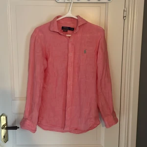 Rosa linneskjorta från Polo Ralph Lauren - Säljer en snygg rosa skjorta från Polo Ralph Lauren i linne. Skjortan har lång ärm, klassisk krage, knäppning framtill och den ikoniska gröna broderade loggan på bröstet. Slim fit-modell som sitter tajt och fräscht. Skjortan är storlek S