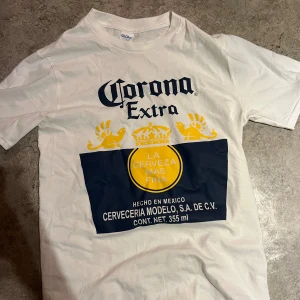 Vit Corona Extra t-shirt - Vit t-shirt med stor Corona Extra-print i blått och gult framtill. Klassisk passform med rund hals och korta ärmar. Tillverkad i mjuk bomull, perfekt för dig som gillar statement-plagg och snygga loggor.
