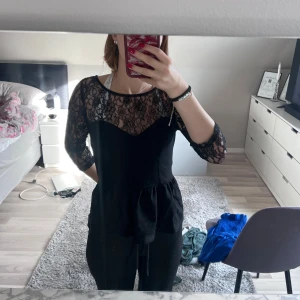 Svart spetsblus  från Guess - Supersnygg svart blus  från Guess med blommig spets upptill och på ärmarna. Blusen  har trekvartsärm, markerad midja och dragkedja i ryggen. Perfekt för dig som vill ha en elegant och trendig look med fina detaljer.