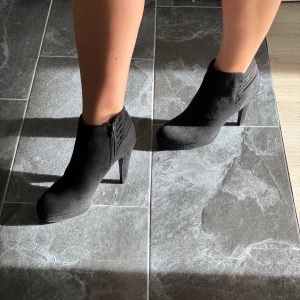 Svarta stövletter med klack - Säljer ett par svarta stövletter med hög klack och rund tå. Skorna har dragkedja på sidan och snygga detaljer vid ankeln. Materialet är mocka-liknande och de har en stilren, elegant look som passar till många outfits.