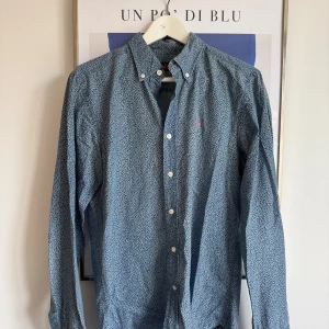 Blå mönstrad skjorta från Morris - Snygg blå skjorta från Morris med smått blommigt mönster och klassisk krage. Skjortan har vita knappar och en broderad rosa detalj på bröstet. Perfekt för dig som gillar stilrena men ändå unika plagg.