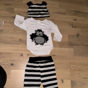 Vit långärmad body med björntryck och byxor till  - Söt vit långärmad body med svart och grått björntryck framtill. Tillhörande mössa och byxor med svart och gråa ränder. Perfekt för dig som gillar gulliga och lekfulla plagg till de minsta. Mjukt material och bekväm passform. 🥰
