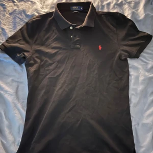 Svart piké från Polo Ralph Lauren - Svart pikétröja från Polo Ralph Lauren i custom fit med klassisk krage och två knappar framtill. Tröjan har det ikoniska röda Polo-logot broderat på bröstet och är tillverkad i 100% bomull för en skön och mjuk känsla. Perfekt för en clean och stilren look.