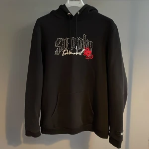 Svart hoodie Supply & Demand M - Svart hoodie från Supply & Demand i storlek M. Snyggt tryck på bröstet med text och röd detalj. Klassisk känguruficka framtill, dragsko i huvan och ribbade muddar. Perfekt för dig som gillar streetwear och vill sticka ut lite extra.