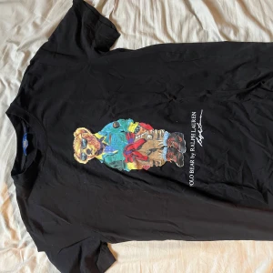 Svart Polo Bear t-shirt från Ralph Lauren - Svart t-shirt från Ralph Lauren med ikoniskt Polo Bear-tryck på bröstet. Klassisk rund hals och korta ärmar. Skön bomullskvalitet och snygg streetstil med färgglad björn i cool outfit framtill.