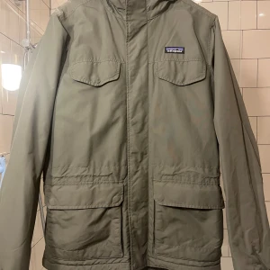 Olivgrön parkajacka från Patagonia - Varm herr retroinspirerad Patagonia M,s Isthmus parka.  Vind och vatten resistent återvunnen nylon med PU beläggning och DWR. Foder i pile-fleece i bål och thermogreen vadderade armar. Avtagbar luva. Justerbar midja. Strl S. Nypris 3200 kr