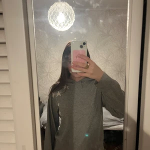 Grå hoodie med stjärnor - Grå hoodie med vita stjärnor på ena axeln och en stor vit stjärna på ryggen. Tröjan har huva och långa ärmar, tillverkad i mjukt bomullsmaterial. Perfekt för en chill och avslappnad stil.