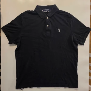Svart pikétröja från U.S. Polo Assn. - Klassisk svart pikétröja från U.S. Polo Assn. med broderad logga på bröstet. Tröjan har krage, korta ärmar och knäppning med tre knappar framtill. Perfekt för en clean och sportig stil. Väldigt snygg och sportig vid biceps                                                     STORLEK M
