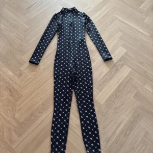 Svart långärmad jumpsuit Chiquelle - Svart långärmad jumpsuit från Chiquelle med vitt rutmönster och logga över hela plagget. Tight passform, hög krage och dragkedja framtill. Tillverkad i stretchigt material som sitter snyggt på kroppen. Perfekt för dig som vill sticka ut med en cool och sportig look.
