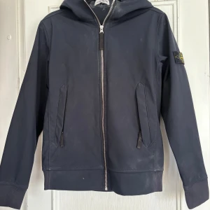 Svart vindjacka från Stone Island Junior - Snygg svart vindjacka från Stone Island Junior med huva och dragkedja framtill. 
