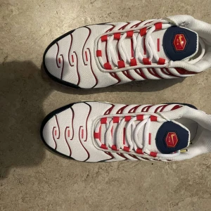 Nike Air Max Plus vita/röda/blå sneakers - Nike Air Max Plus sneakers i vitt mesh med röda och blå detaljer samt gula accenter. Snygg vågig design på ovandelen, blå sula med synlig Air-enhet och Tn Air-logga på hälen. Skorna har snörning och en sportig vibe, perfekt för dig som vill sticka ut.