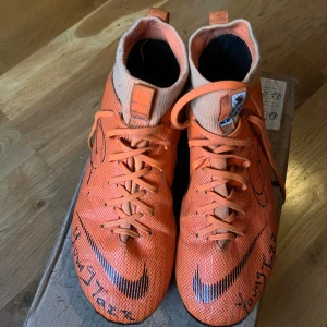 Nike Mercurial orange fotbollsskor - Säljer ett par grymma Nike Mercurial fotbollsskor i orange med svarta detaljer och snörning. Skorna har högre strumpliknande skaft för extra stöd och är tillverkade i syntetmaterial. Perfekta för dig som vill sticka ut på planen!