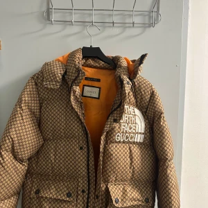 Gucci x The North Face pufferjacka  - Säljer en fet pufferjacka från Gucci x The North Face i beige med brunt monogrammönster och orange insida. Jackan har huva, stora fickor och broderad logga på bröstet. Perfekt statement-plagg för dig som vill sticka ut. Storlek är S med kan bassar M