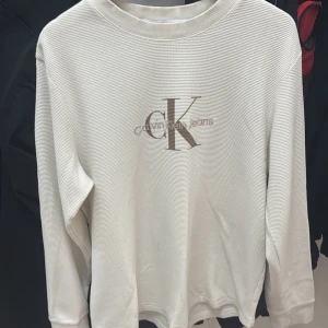Vit långärmad tröja Calvin Klein Jeans - Vit långärmad tröja från Calvin Klein Jeans med diskret ribbad struktur och stort CK-logotyptryck framtill. Tröjan har rund halsringning och är tillverkad i mjuk bomull för en clean och stilren look.det lila på bilden är min kamera som är sönder 