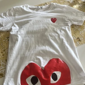 Vit Comme des Garçons Play t-shirt - Vit t-shirt från Comme des Garçons Play med det ikoniska röda hjärtat med ögon broderat på bröstet och ett stort tryckt hjärta längst ner. Klassisk rund hals och korta ärmar. Tillverkad i mjuk bomull för en skön känsla. Den är för liten för mig. Jag är 185 cm men den är jättefin!