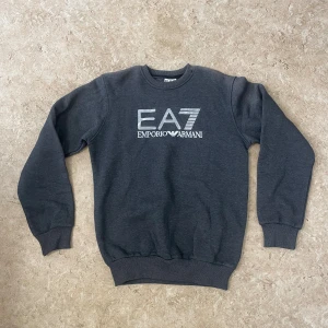Mörkgrå EA7 Emporio Armani sweatshirt - Snygg mörkgrå sweatshirt från EA7 Emporio Armani med stort logotryck i vitt på bröstet. Tröjan har rund hals, ribbade muddar och är tillverkad i mjuk bomullsblandning. Perfekt för en stilren och avslappnad look.