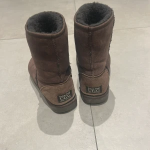Bruna UGG boots i mocka - Mysiga bruna UGG boots från Urban Trail, fodrade med mjuk päls och tillverkade i mocka. Klassisk rund tå och platt sula, perfekta för kalla dagar. Märke på hälen och enkel slip-on modell. Nästintill aldrig använda! Perfekta nu inför höst/vinter. Öppen för att diskutera pris.