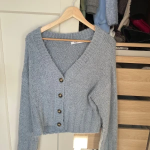 Ljusgrå stickad kofta från NA-KD - Supermysig ljusgrå stickad kofta från NA-KD med v-ringning och fyra snygga knappar framtill. Koftan är croppad och har långa ärmar samt bred ribbad kant nedtill och vid ärmsluten. Perfekt för lager-på-lager under kyliga dagar.