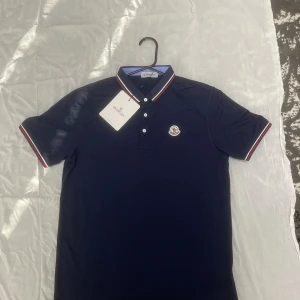 Moncler Pike  - 💎 Moncler Piké – Helt Ny med Tag 💎 🔥 Exklusiv Moncler piké i klassisk marinblå färg – perfekt blandning av stil och lyx! Storlek M 👕 Aldrig använd – helt ny med tag och scan ✅ 🔹 Detaljer: 	•	Ikoniskt Moncler-emblem på bröstet 🇫🇷 	•	Snygga ränder i rött, vitt och blått på krage & ärmslut 	•	Högkvalitativ bomull som känns lyxig och bekväm 	•	Passar både till vardags och finare tillfällen 📦 Skickas samma dag om köpet görs innan 20:00! 💰 Riktigt fin piké för dig som vill ha stil och kvalité