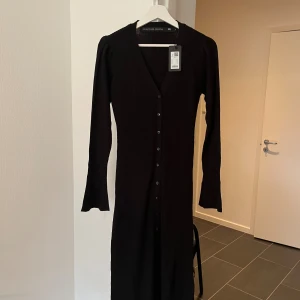 Svart ribbad klännning/cardigan från Lager 157 - Oanvänd svart lång ribbstickad klänning/cardigan från The Black Label Collection. Klänningen har v-ringning, långa ärmar och knappar hela vägen framtill. Tillverkad i 100% akryl och har en slim passform som framhäver siluetten. Perfekt för dig som gillar stilrena och tidlösa plagg.