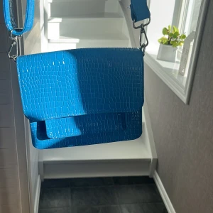 Blå axelväska i krokopräglad syntet - Cool blå axelväska med bred axelrem och krokopräglad yta. Väskan har en fyrkantig form och stängs med lock. Perfekt för dig som vill sticka ut med en färgstark accessoar. Materialet är glansig syntet och väskan har silvriga metalldetaljer.