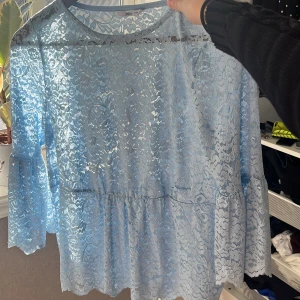 Blå spetsblus från Ellos - Säljer en ljusblå blus från Ellos i transparent spets med blommigt mönster. Blusen har trekvartsärmar med volang och rund halsringning. Perfekt för dig som vill ha en romantisk och trendig look.