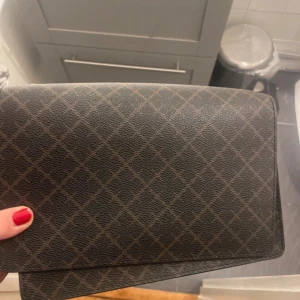 Malene Birger väska  - Snygg axelväska från By Malene Birger i mörkbrunt och svart med rutigt mönster. Väskan har bred axelrem med gulddetaljer och flera fack för smart organisering. Tillverkad i skinn  material och perfekt för dig som vill ha en trendig och stilren look.