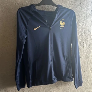 Nike FFF hoodie i marinblå Dri-FIT  - Marinblå hoodie från Nike med FFF-logga och guldiga detaljer. Tillverkad i Dri-FIT polyester, hel dragkedja framtill, huva och fickor. Perfekt för fotboll eller annat.
