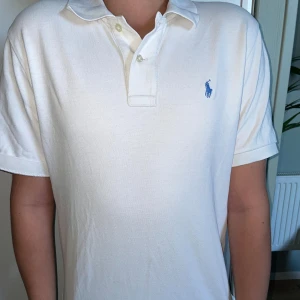 Vit pikétröja från Ralph Lauren - Vit pikétröja från Ralph Lauren med klassisk krage och två knappar framtill. Tröjan har korta ärmar och den ikoniska blå broderade loggan på bröstet. Tillverkad i mjuk bomull som är skön att ha på sig.