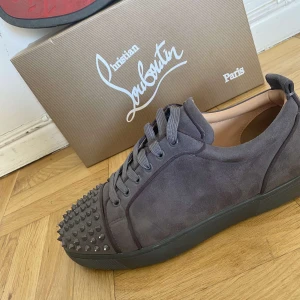 Grå Christian Louboutin sneakers med nitar - Snygga grå sneakers från Christian Louboutin i mocka med coola nitar på tån och klassisk röd sula. Skorna har snörning och en rund tå, vilket ger dem en edgy vibe. Perfekta för dig som vill sticka ut med exklusiva detaljer och lyxig känsla. Använda! Storlek 43