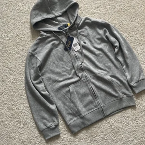 Grå hoodie från Polo Ralph Lauren - Snygg grå hoodie från Polo Ralph Lauren med dragkedja framtill, huva med snörning och klassisk logga broderad på bröstet. Tillverkad i mjuk bomull och har två stora fickor framtill. Perfekt för en chill och stilren look.