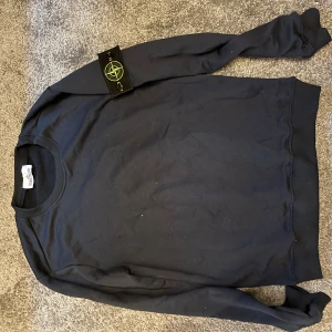 Mörkblå sweatshirt från Stone Island - Säljer en mörkblå sweatshirt från Stone Island med klassisk rund hals och ikonisk patch på vänster ärm. Tröjan är i mjuk bomull och har ribbade muddar vid ärmslut och nederkant. Perfekt för dig som gillar streetwear och stilrena plagg.