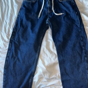 Blå district fortysix med snörning - Säljer ett par mörkblå jeansbyxor från District 46 med vit snörning i midjan och raka ben. Klassisk femficksmodell med broderad logga på bakfickan. Perfekta för dig som gillar en avslappnad och stilren look.