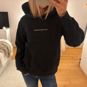 Svart hoodie från WeSC - Svart hoodie från WeSC med vit broderad text 'WeAretheSuperlativeConspiracy' på bröstet. Klassisk känguruficka, huva med snörning och mjukt bomullsmaterial. Perfekt för chill dagar och streetstyle.