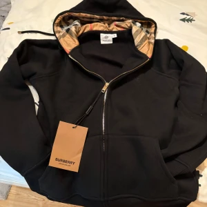 Svart hoodie från Burberry med dragkedja -  Storlek S !!!! Svart hoodie från Burberry med klassiskt rutigt foder i huvan och guldfärgad dragkedja. Tröjan har broderad logga på bröstet, känguruficka och snörning i huvan. Tillverkad i mjuk bomull med lite elastan för extra komfort.