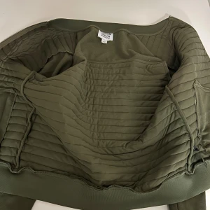 Olivgrön quiltad bomberjacka Burton London - Snygg olivgrön bomberjacka från Burton London i storlek L. Jackan har quiltade paneler, ribbade muddar och silverfärgad dragkedja. Materialet är polyester och jackan har en loose passform med långärmade ärmar. Perfekt för dig som gillar stilren och urban look.