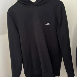 Svart hoodie från A.P.C. i bomull - Svart hoodie från A.P.C. med minimalistisk vit logga på bröstet. Klassisk modell med huva och långa ärmar, tillverkad i mjuk bomull. Perfekt för en clean och stilren look. Sann i storleken och passar därav medium. Fint skick. Kom i DM för bud